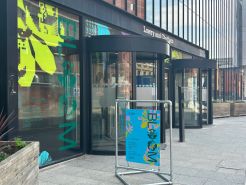 An image of the outside of the Lowry Building - placed before the revolving doors is a standalone sign saying 'Bloom' (the name of the degree show). On the windows of the front of the building are huge graphic flowers and the title 'BLOOM'.