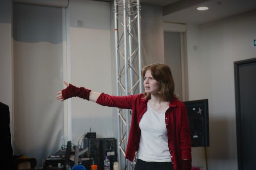 Person in a red cardigan and white top stands indoors with one arm outstretched during a presentation or performance. Behind the person are lighting trusses, audiovisual equipment and a monitor in a modern event or studio space.