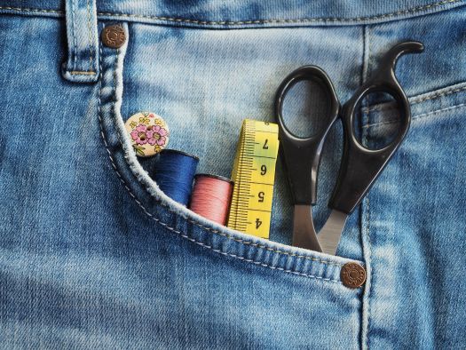 Close-up of a blue denim jeans pocket containing sewing supplies, including black-handled scissors, a yellow measuring tape, two spools of thread (blue and pink), and a floral-patterned button. The image highlights clothing repair, sewing, or upcycling.