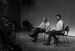 The award winning playwright and former BBC script editor Ray Grewal sits on a chair on a stage. He's flanked by a man to the left of him.